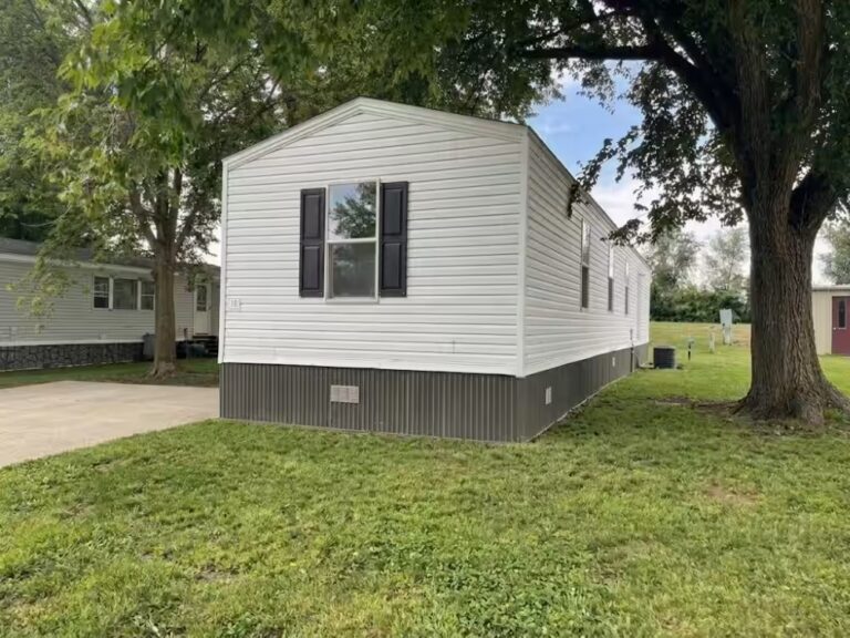 White mobile home with black shutters in a grassy yard surrounded by large trees