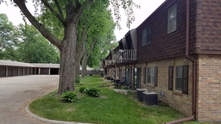 Brick apartment building with balconies and trees beside a lawn and garage parking lot.