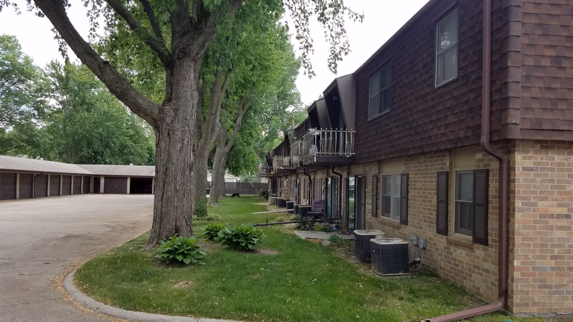 Brick apartment building with balconies and trees beside a lawn and garage parking lot.