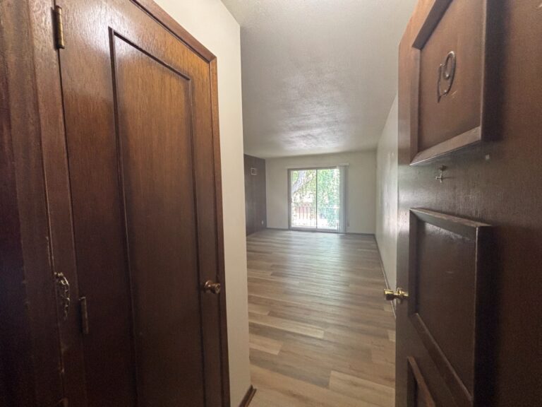 Open wooden door numbered 19 revealing empty apartment room with wood floors and sliding glass door