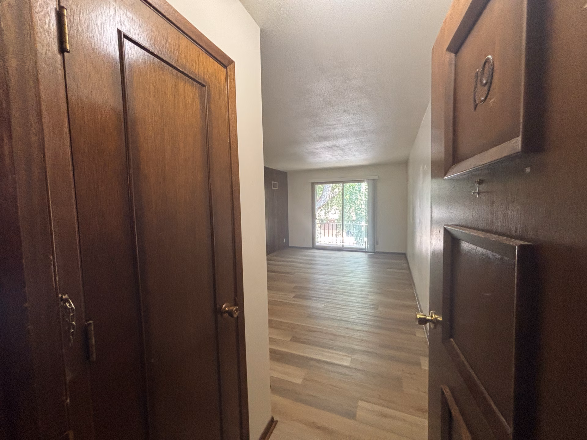 Open wooden door numbered 19 revealing empty apartment room with wood floors and sliding glass door
