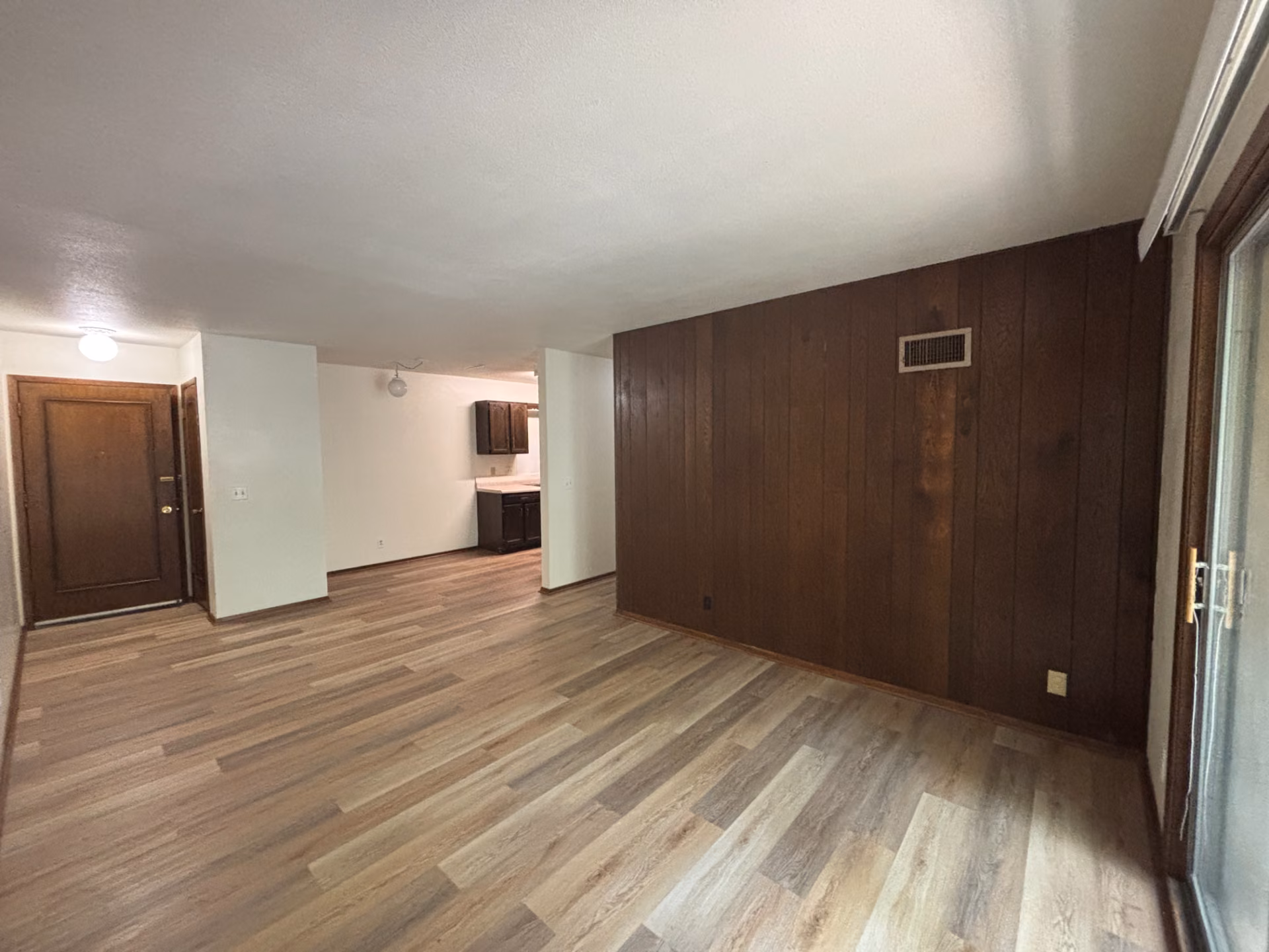 Empty living room with dark wood panel accent wall, wood-look flooring, and kitchen visible behind.