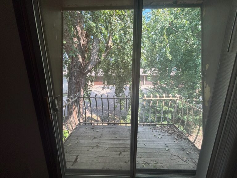 Weathered wooden balcony with metal railing surrounded by trees, viewed through sliding glass door.