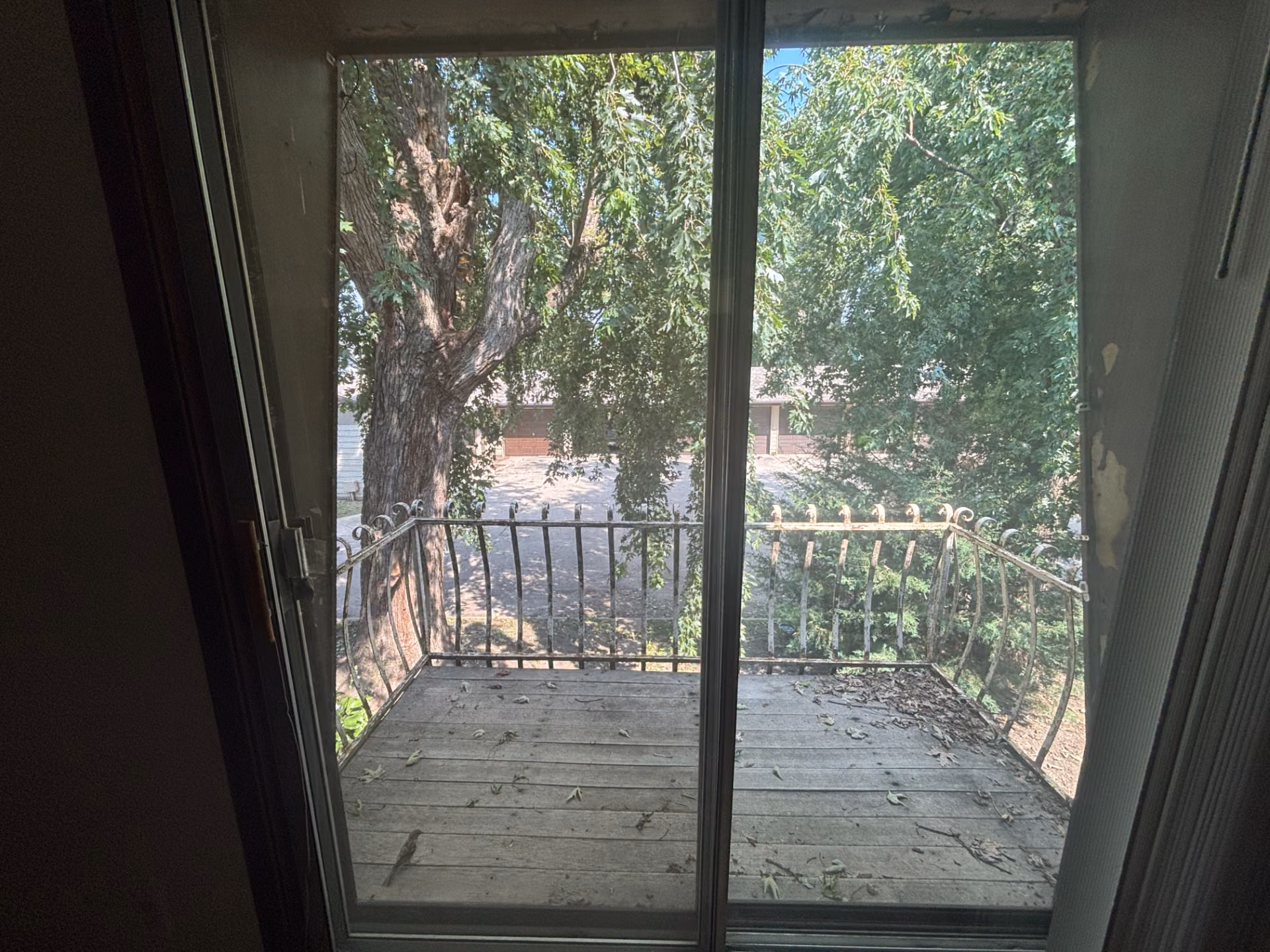 Weathered wooden balcony with metal railing surrounded by trees, viewed through sliding glass door.