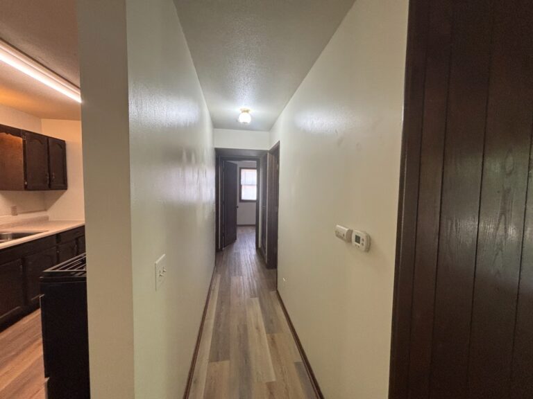 Home hallway with wood flooring, cream walls, dark wood paneling, and kitchen view on left.