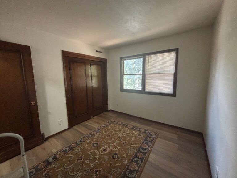 Empty room with wood floors, an ornate area rug, wooden closet doors, and a window with blinds