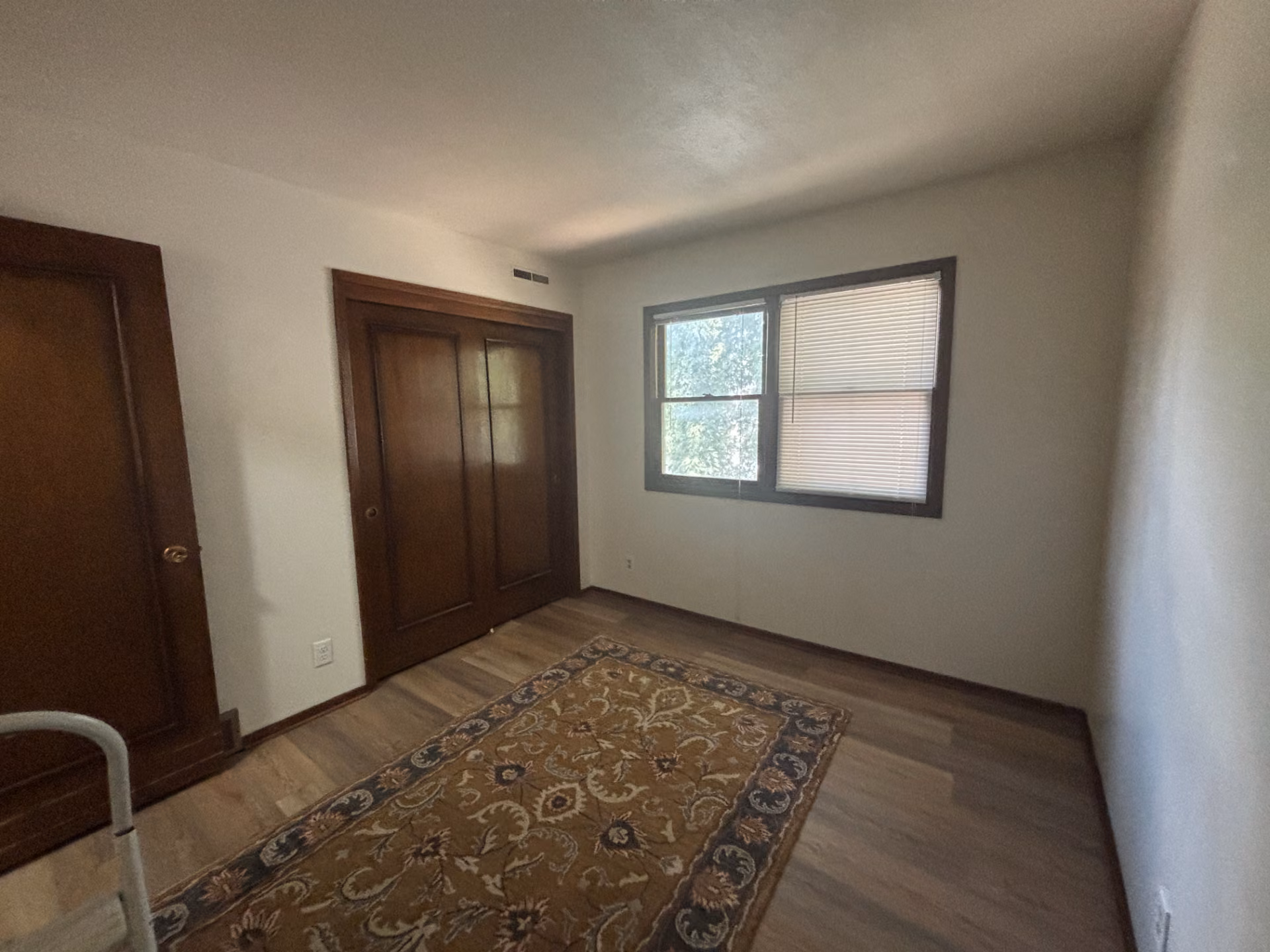 Empty room with wood floors, an ornate area rug, wooden closet doors, and a window with blinds