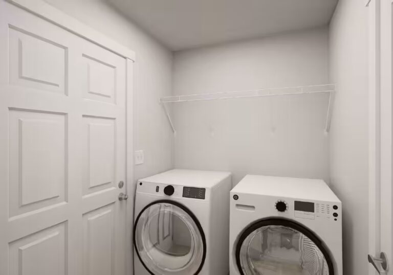 Laundry room with white GE washer and dryer beneath white shelf, beside white paneled door.