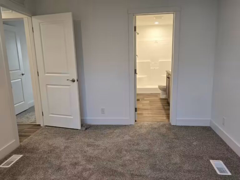Empty room with gray carpet, white door ajar, and open doorway to a bathroom with toilet and tub.