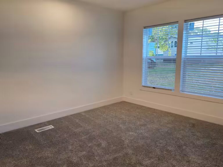 Empty room with gray carpet and two windows with white blinds overlooking a green yard