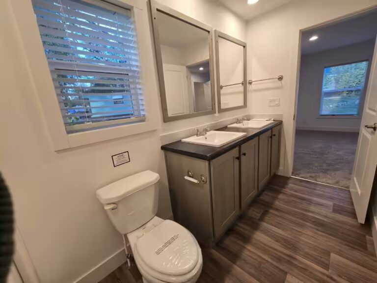 Bathroom with white toilet, double sink vanity, two mirrors, wood-look flooring, and open doorway.