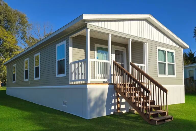 Gray manufactured home with covered porch and wooden stairs on a green lawn under blue sky.