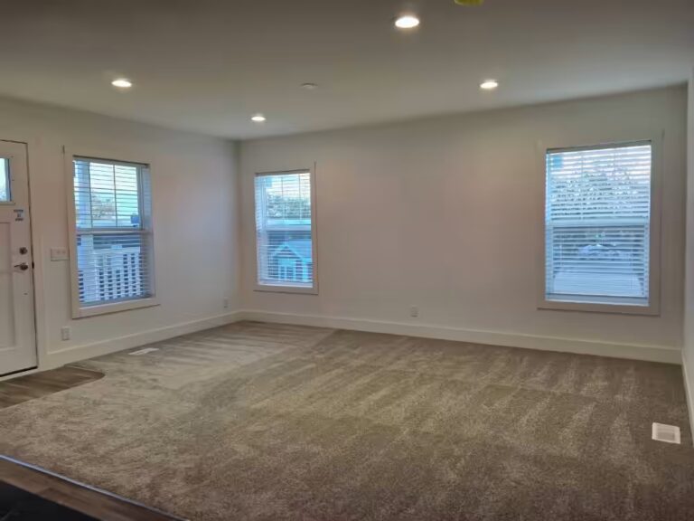 Empty room with beige carpet, three windows with blinds, white walls, and a white entry door.