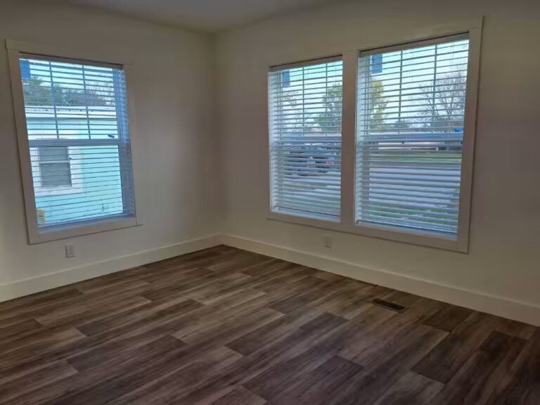 Empty room with wood-look flooring and windows with blinds overlooking houses