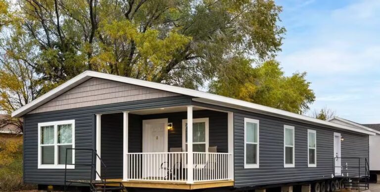 Dark gray single-wide mobile home with covered white porch and railing, set among autumn trees.