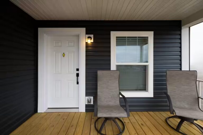 Two gray patio chairs on a wooden porch beside a white door and window of a house with dark siding.