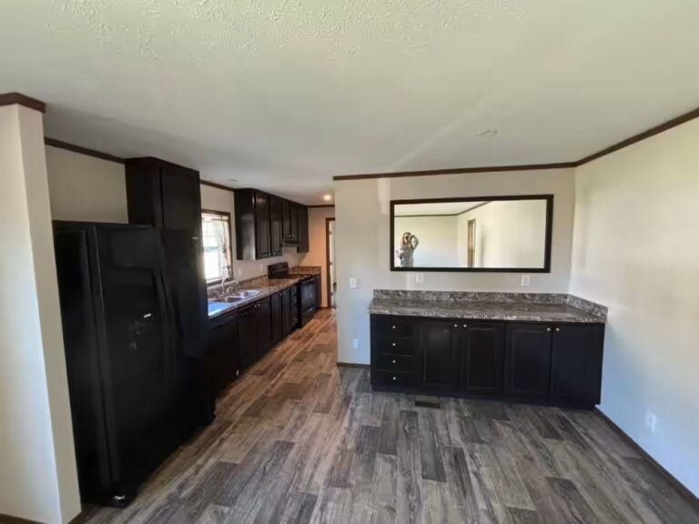 Empty kitchen with dark cabinets, black fridge, granite counters, wood floors, and wall mirror