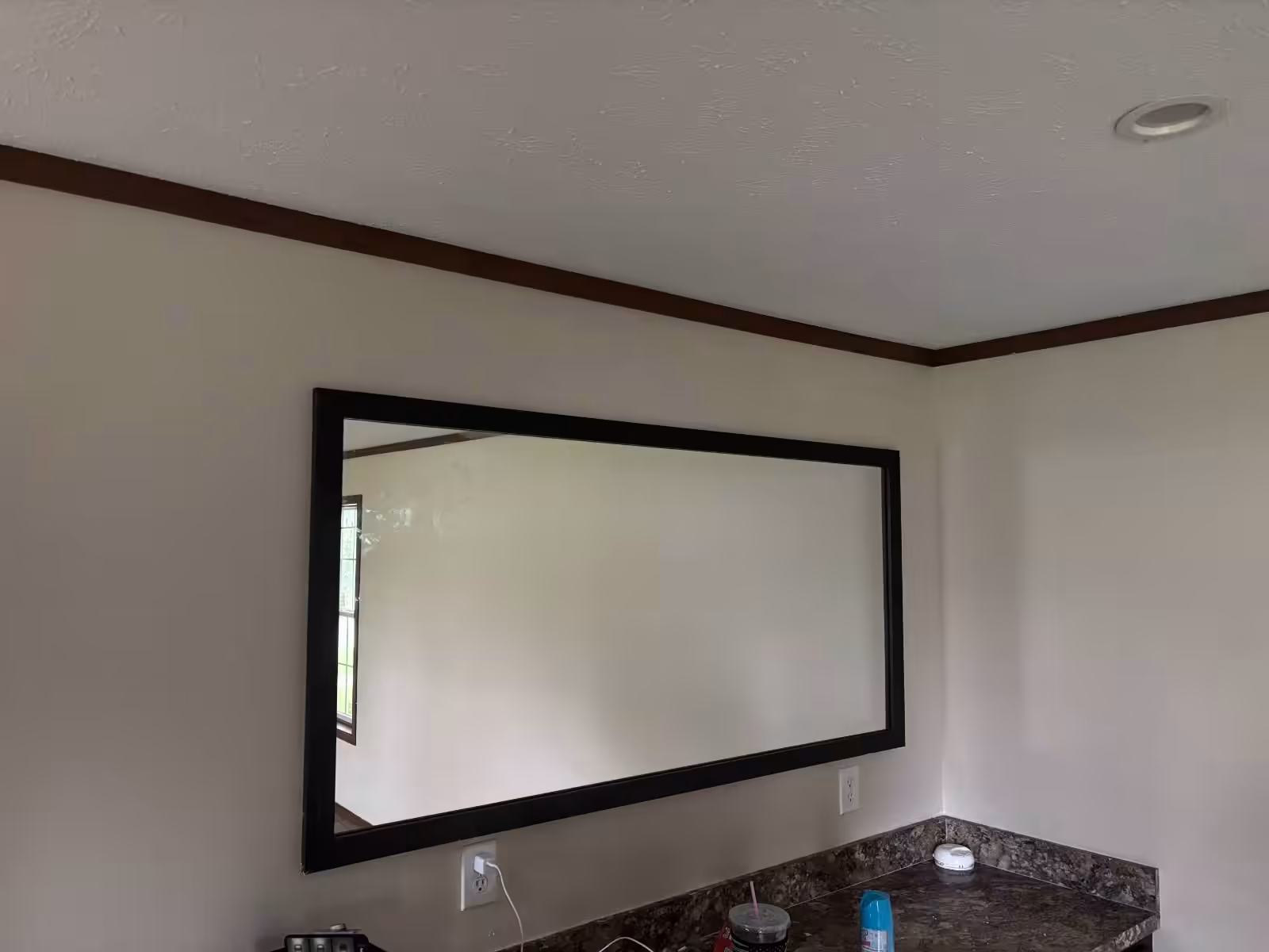 Large rectangular mirror with black frame mounted on beige wall above granite countertop