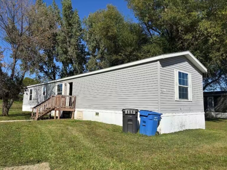 Gray single-wide mobile home with wooden deck stairs, trees behind and trash bins in front yard