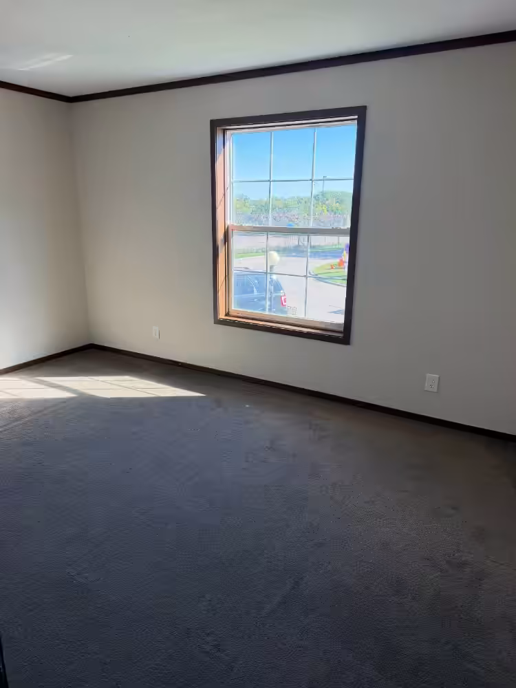 Empty room with beige walls, gray carpet, and a window overlooking a parking area and trees