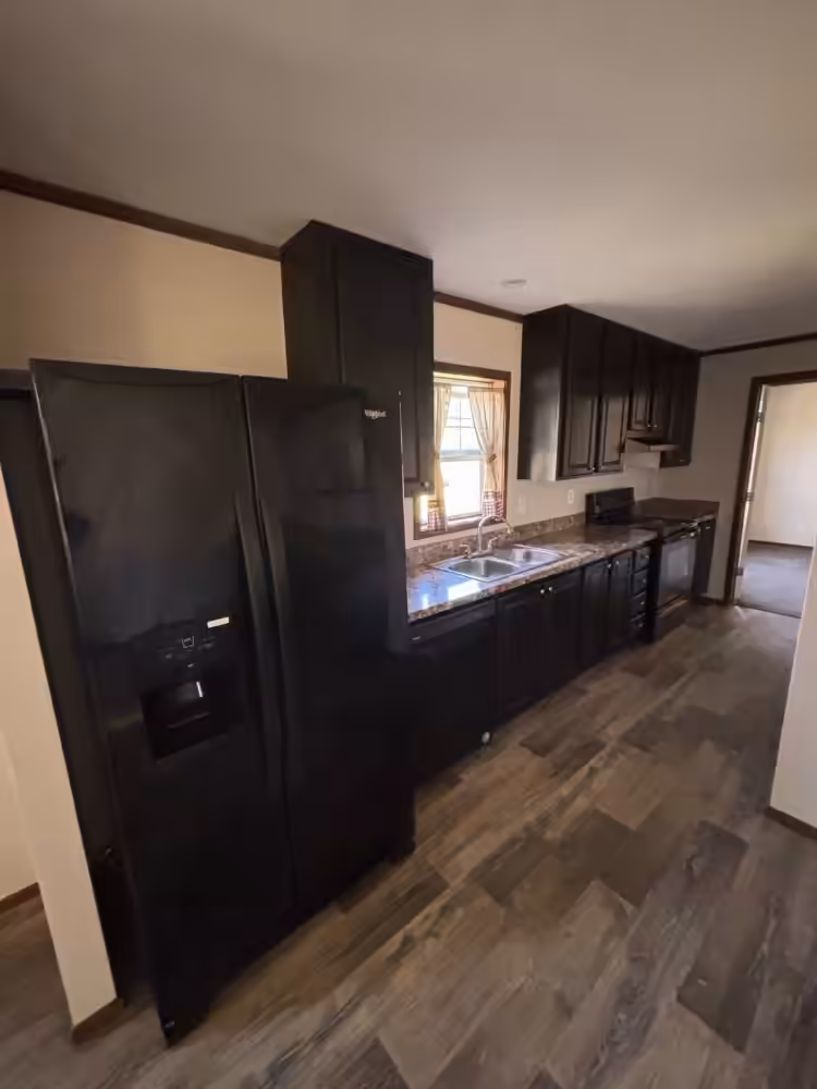 Kitchen with black refrigerator, dark cabinets, sink under window, and wood-look flooring.