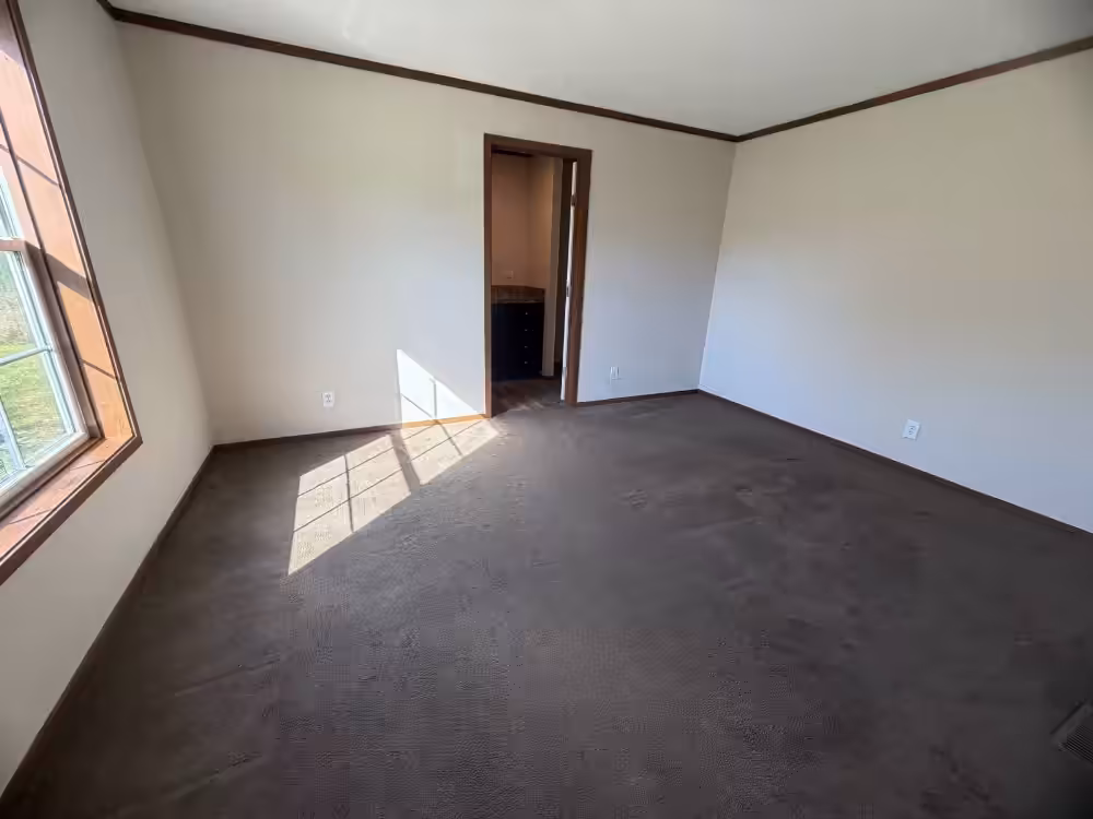 Empty room with white walls, brown carpet, a window on the left, and an open doorway to another room.