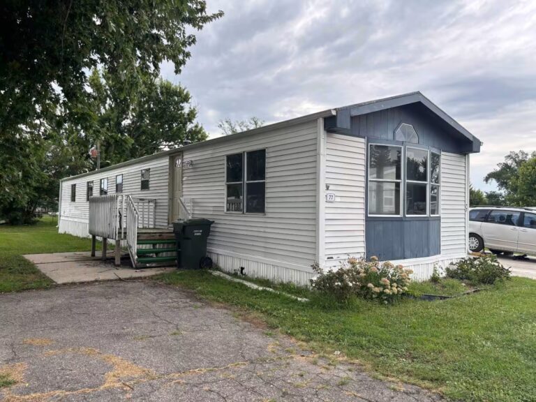 White single-wide mobile home with blue trim, wooden entry ramp, marked 72, under a cloudy sky.