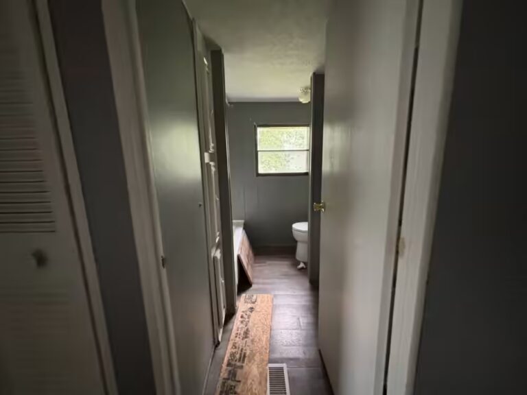 Narrow home hallway with plywood board on floor, window and toilet visible at far end