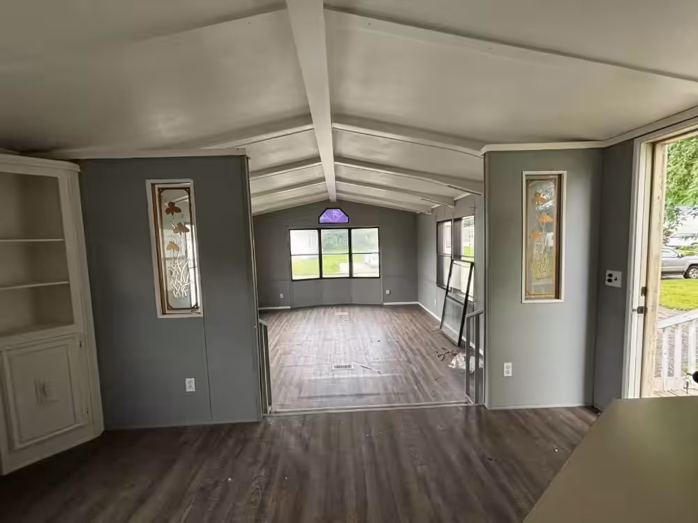 Empty room with vaulted ceiling, wood floors, gray walls, and windows facing a green yard.