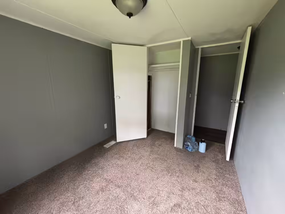 Empty room with gray walls, beige carpet, open white door, closet, and ceiling light fixture