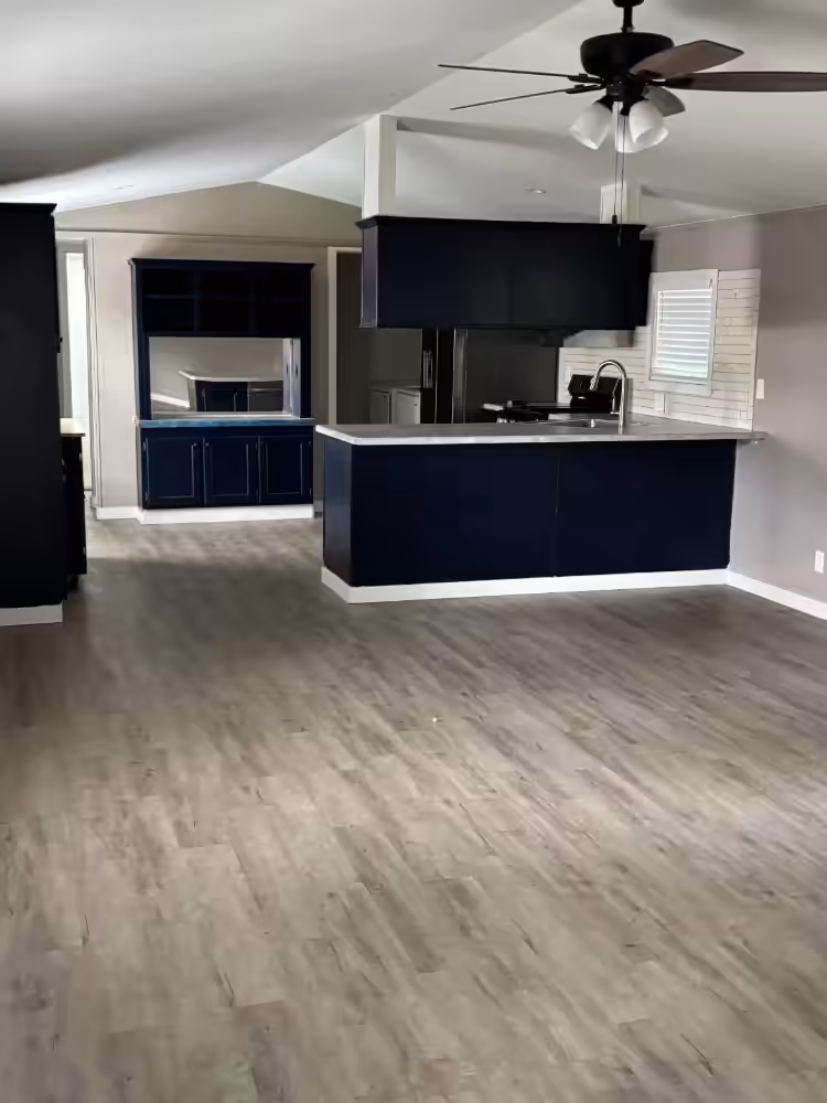 Empty open-plan kitchen with navy blue island and cabinets, gray wood-look flooring, and ceiling fan.
