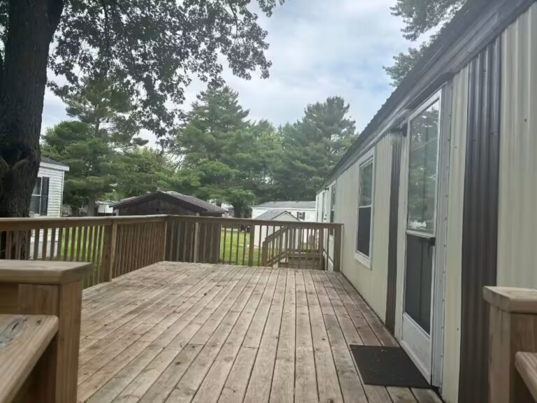 Wooden deck beside white mobile home with screened door, railings, trees, and sheds in background.