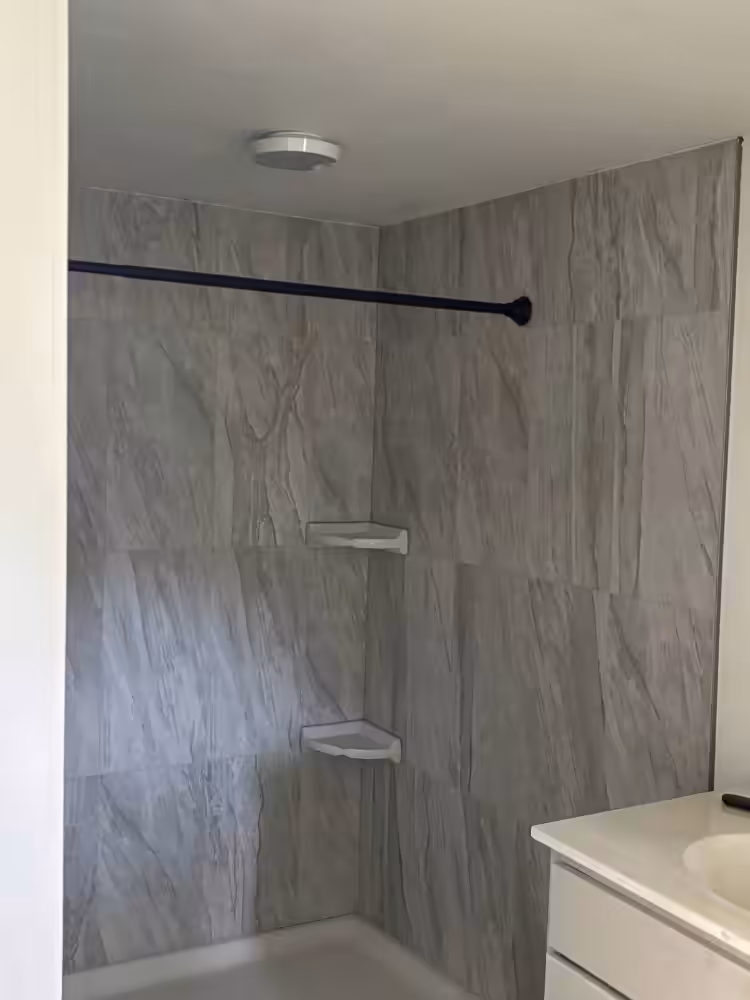 Shower with gray marble-look walls, black curtain rod, corner shelves, and white vanity sink.