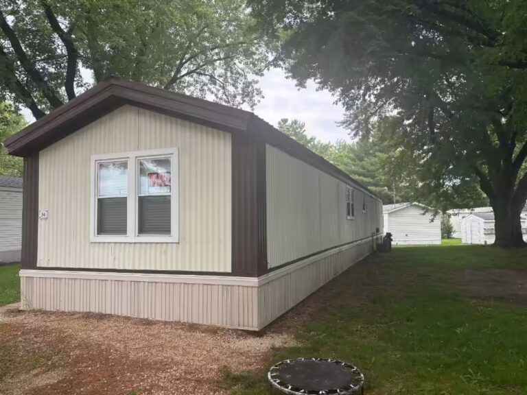 Mobile home with white siding and dark brown trim, trees and grassy yard surrounding it.