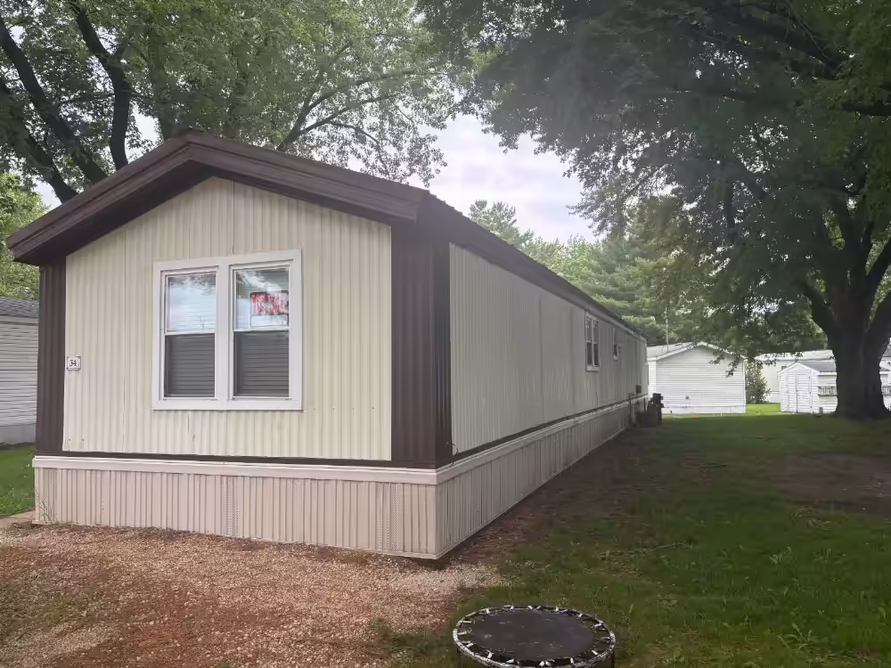 Mobile home with white siding and dark brown trim, trees and grassy yard surrounding it.