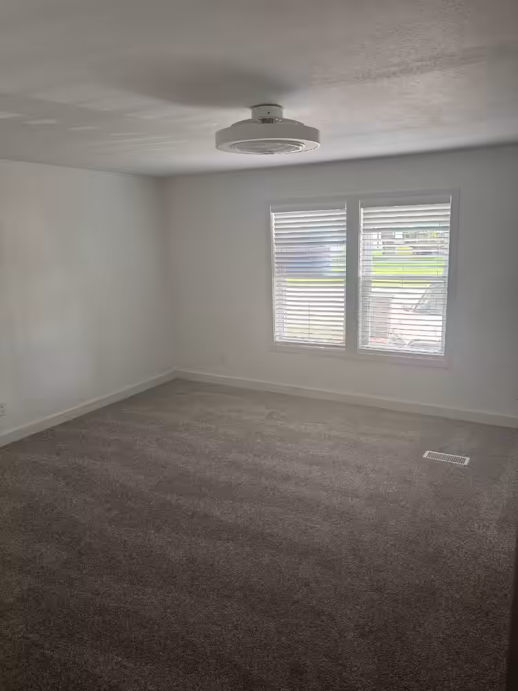 Empty room with white walls, gray carpet, ceiling light, and two windows with white blinds.