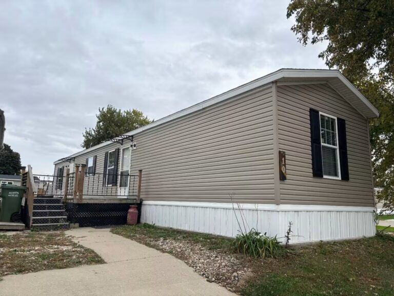 Beige mobile home with wooden deck and stairs, under cloudy sky, address plaque showing 10.