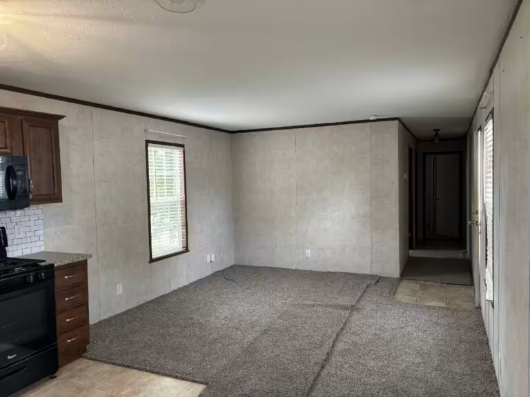 Empty room with gray carpet, beige walls, and kitchen area with dark wood cabinets and black stove